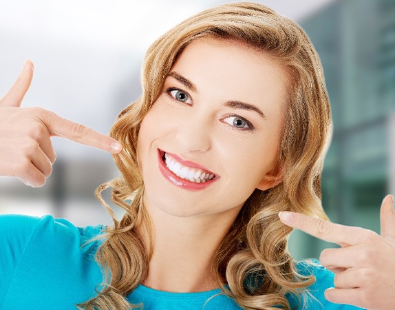 Woman in teal shirt pointing to her smile with both hands