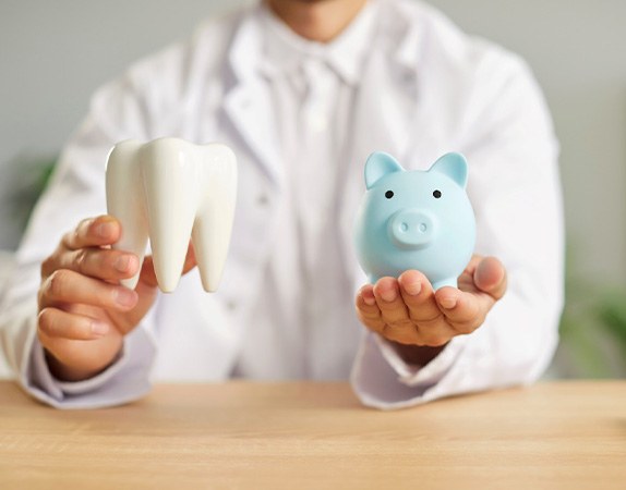 Neck-down view of man in white coat holding large model tooth and blue piggy bank